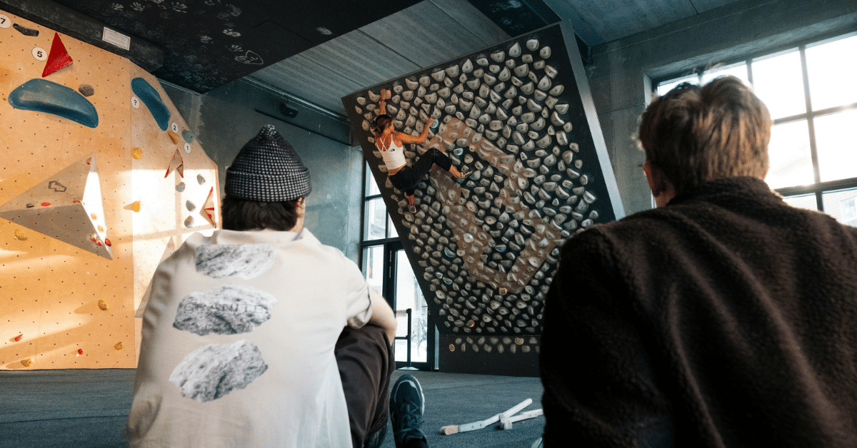 The Climber’s Guide to Hand, Wrist, and Finger Injuries: Why Rest Alone Doesn’t Cut It Two climbers sit on the floor of an indoor climbing gym, watching another climber tackle a steep overhanging bouldering wall.